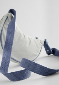 Borsa in tessuto grigio chiaro con una forma curvata, caratterizzata da una tracolla e bordi blu testurizzati. La tracolla è dotata di una fibbia in plastica sicura.