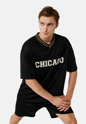 Junger Mann in schwarzem "CHICAGO" Trikot und Shorts, dehnt die Beine mit verschränkten Händen über dem Knie, schaut nach links.