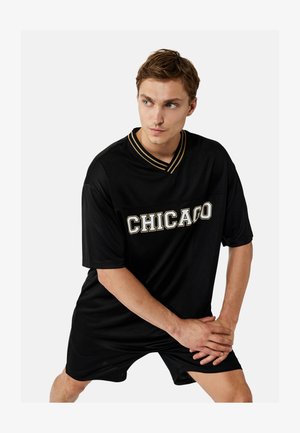 Junger Mann in schwarzem "CHICAGO" Trikot und Shorts, dehnt die Beine mit verschränkten Händen über dem Knie, schaut nach links.