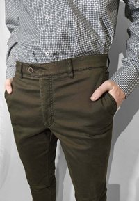 Pantaloni verde scuro con una texture liscia, dotati di tasche frontali e chiusura con bottone, abbinati a una camicia aderente con motivo blu e bianco.