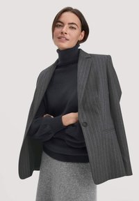 Grau gepinsterter Blazer mit Schalkragen, getragen über einem schwarzen Rollkragenpulli. Weiche Textur und strukturiertes Design. Ein hellgraßer Rock rundet das Outfit ab.