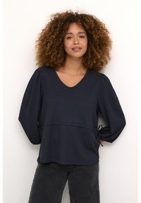 Haut texturé bleu marine avec un décolleté en V, des manches bouffantes et des liens réglables à la taille. Associé à un jean sombre.