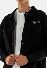 Chemise à boutons en velours côtelé noire avec deux poches avant et logo brodé blanc "Project Paris", portée sur un t-shirt blanc.