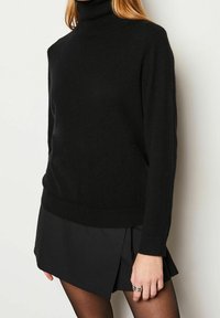 Pull à col roulé noir en tissu tricoté doux et texturé, à manches longues et coupe légèrement ample, assorti à une jupe plissée noire.