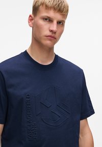 Tumši zila kokvilnas T-krekls ar teksturētu logo tumšākā zila tonī, ar apaļu kakla izgriezumu, īsām piedurknēm un sānu šuvi, lai izveidotu pieguļošu siluetu.