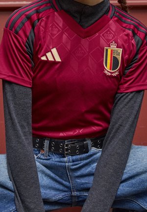 Červený fotbalový dres s černými akcenty, diamantovým vzorem, zlatým logem Adidas a erbem Belgie. Nalezeno na šedém tričku s dlouhým rukávem, v kombinaci s modrými džíny.