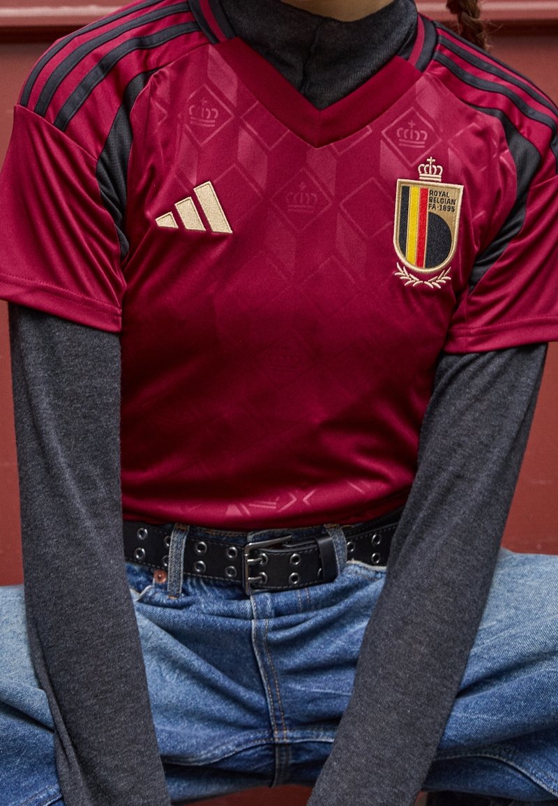 Červený fotbalový dres s černými akcenty, diamantovým vzorem, zlatým logem Adidas a erbem Belgie. Nalezeno na šedém tričku s dlouhým rukávem, v kombinaci s modrými džíny.