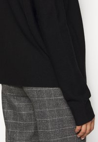 Pulover negru tricotat, cu o croială relaxată și mâneci lungi, asortat cu pantaloni gri din țesătură tartan, cu un model în carouri.