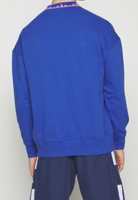 Personne portant un sweat-shirt bleu uni avec un détail de col rose à motif, associé à un pantalon bleu marine foncé à bandes blanches, vue de dos.