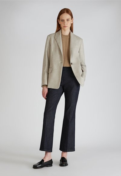 Blazer grigio chiaro su un maglione di cotone beige, abbinato a pantaloni a campana in denim scuro e mocassini neri. Sfondo neutro, modello in piedi.