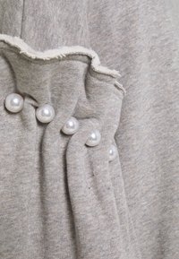 Tela gris con detalle de costura rizada y cinco cuentas de perla blanca decorativas espaciadas uniformemente a lo largo del pliegue.