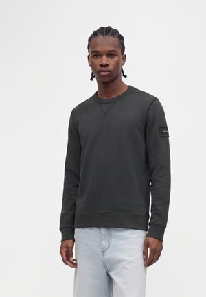 Jeune homme aux cheveux tressés portant un sweat-shirt à manches longues foncé et un jean bleu clair, debout devant un fond clair uni.