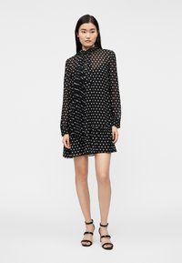 Γυναίκα που φοράει μαύρο διάφανο φόρεμα με μακριά μανίκια και λευκές πουά, με φουλάρι δέσιμο στον λαιμό και μαύρα πέδιλα με ψηλά τακούνια και λουράκια, όρθια.