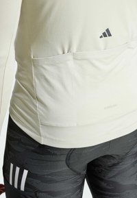 Jasnobeżowa, długi rękaw, sportowa bluzka o gładkiej fakturze, z kieszonką z tyłu i czarnym logo. Połączona ze wzorzystymi czarnymi legginsami.