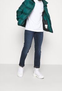 Chaqueta acolchada verde azulado con un acabado brillante, camiseta blanca, vaqueros azules con los puños remangados y zapatillas blancas. Conjunto casual con texturas superpuestas.