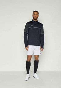 Námornícky športový dlhý rukáv s bielymi akcentmi, skombinovaný s bielymi šortkami a čiernymi podkolienkami. Obsahuje branding Under Armour.