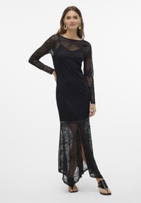 Svart spetsklänning med långa ärmar, med geometriska mönster, en figursydd bodice och en semi-transparent kjol med en hög slits. Svarta sandaler.