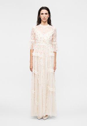 Femeie purtând o rochie albă transparentă până în pământ cu broderii florale, mâneci trei sferturi și detalii cu volane, stând pe un fundal simplu.