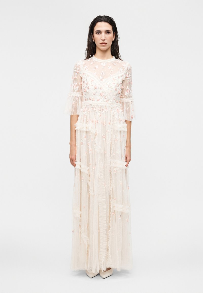 Γυναίκα που φοράει λευκό αέρινο φόρεμα μέχρι το έδαφος με κεντήματα λουλουδιών, μανίκια ως τον αγκώνα και λεπτομέρειες με βολάν, στέκεται μπροστά σε μία απλή φόντο.