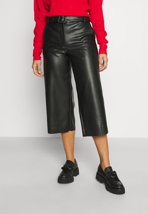 Vrouw draagt zwarte leren cropped broek met wijde pijpen, zwarte riem, zwarte chunky loafers en een rode top met lange mouwen, staande op een grijze vloer.