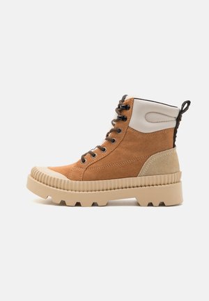 Bruine suède enkellaarzen met een witte stoffen kraag, dikke beige zool en zwarte veters. Voorzien van geribbelde zoolstructuur en contrasterende accenten.