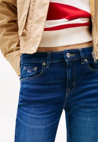 Blå jeans av högmidjad denim med kopparfärgade detaljer, fem fickor och en liten logotyp på framfickan. Slät textur.