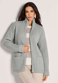 Eine Frau trägt eine hellgraue, gerippte Zip-Up-Strickjacke über einem beige-weißen gestreiften Pullover und hält den Reißverschluss der Strickjacke in der Nähe ihrer Taille.