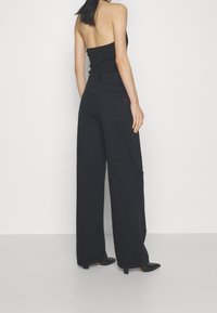 Black wide-leg jeans with high waist, featuring a red logo tag. Smooth denim texture, paired with a fitted black top and pointed black heels.