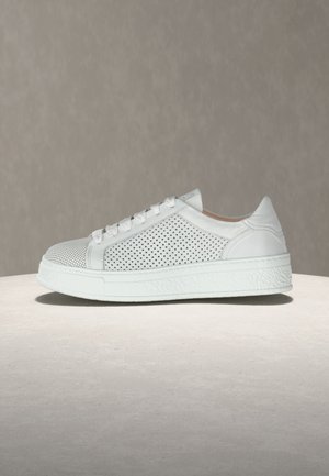 Witte lage sneaker met geperforeerde zijkanten en witte veters, getoond op een licht stenen oppervlak tegen een grijze achtergrond.