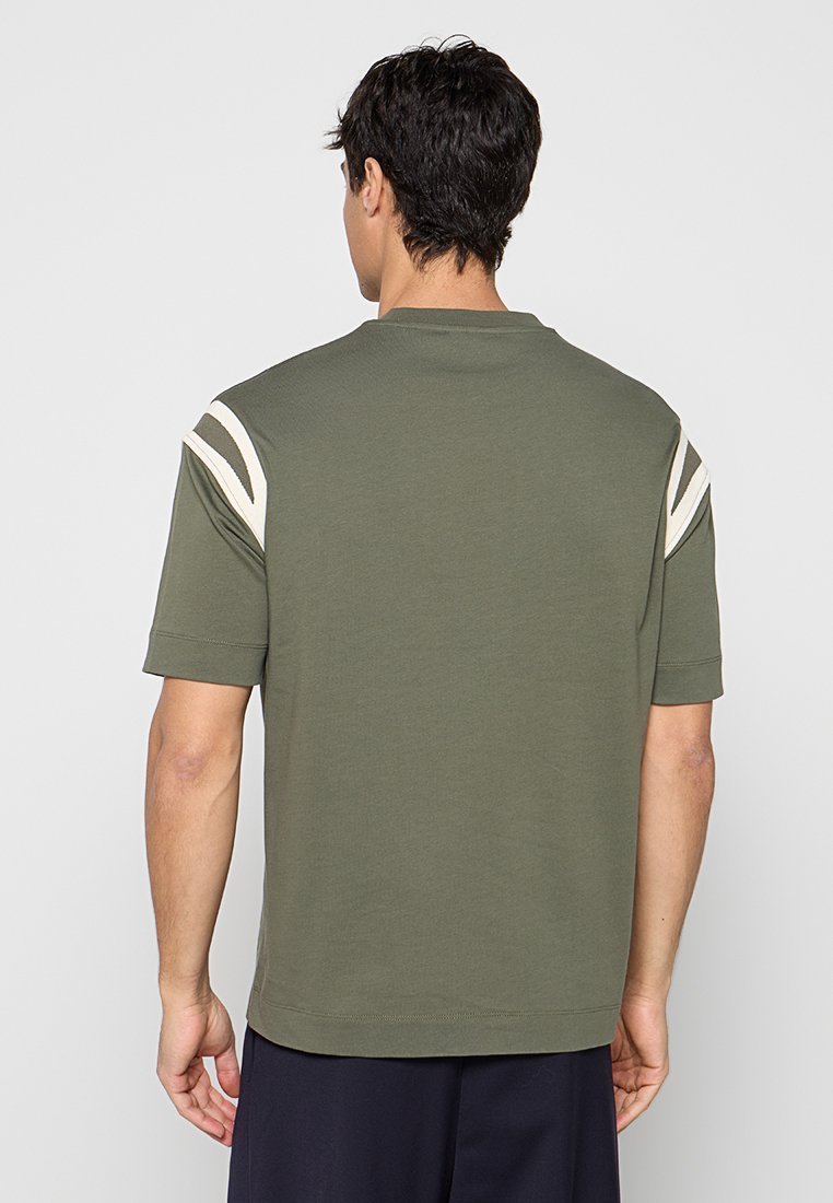 Hombre de espaldas que lleva una camisa verde oliva de manga corta con rayas blancas en los hombros y pantalones oscuros sobre un fondo liso.
