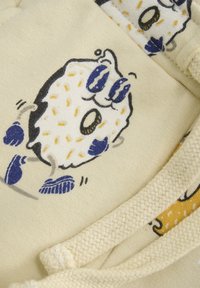 Baumwollstoff in cremigem Beige mit einem Cartoon-Style Donut-Druck in Schwarz, Weiß und Blau. Zugband-Detail mit strukturiertem Gewebe.