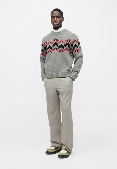 Γκρι πλεκτό πουλόβερ με γεωμετρικό σχέδιο σε κόκκινο, μαύρο και γκρι, συνδυασμένο με ανοιχτόχρωμες γκρι παντελόνες και πολύχρωπα παπούτσια.