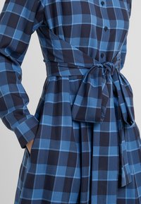 Robe à carreaux bleue et bleu foncé avec manches longues, devant boutonné, ceinture nouée à la taille et main placée dans une poche latérale.
