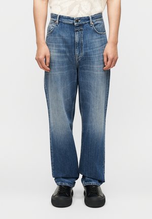 Person wearing loose-fit blue faded jeans and black sneakers, standing against a plain white background.