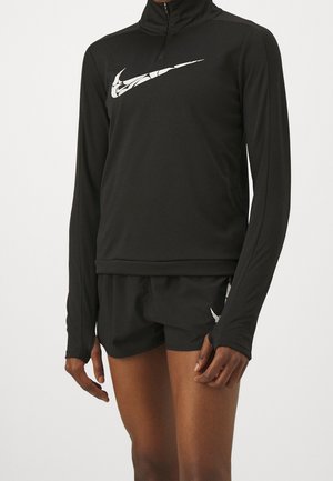 Person trägt schwarzen langärmligen Sporttop mit weißem Nike-Swoosh und passende schwarze Shorts mit weißem Nike-Logo am linken Oberschenkel.