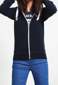 Personne ajustant la capuche d'un sweat à capuche zippé bleu marine sur un t-shirt graphique bleu marine, portant un jean en denim bleu sur fond uni.