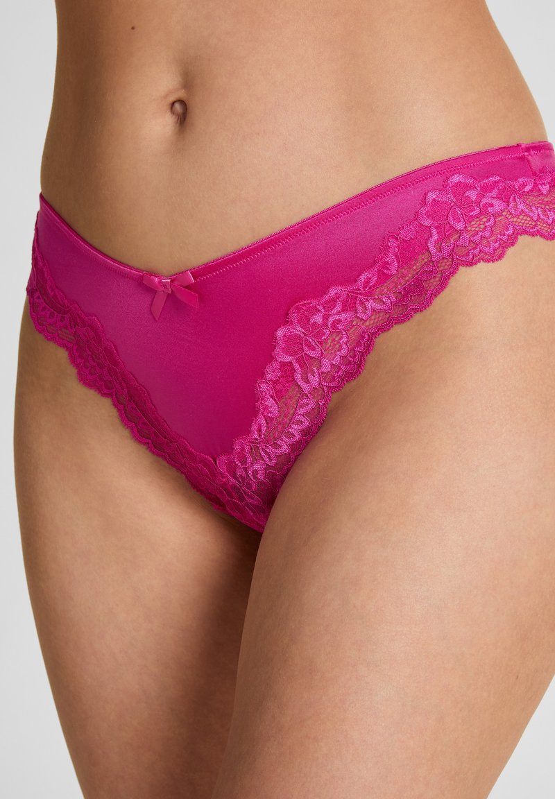 Vrouw draagt felroze satijnen bikini-ondergoed met kanten rand en een kleine strik aan de voorkant in het midden.