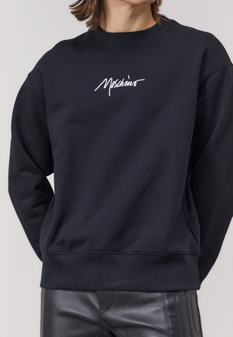 Sweat-shirt noir en tissu doux, avec un col rond et des manches longues. Orné d'un logo blanc brodé sur la poitrine.