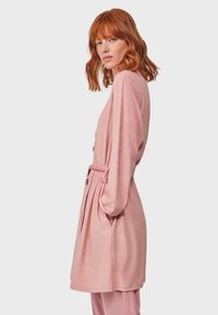 Accappatoio lungo rosa chiaro con vita a cintura, dettagli con bottoni e tasche laterali. Realizzato in tessuto morbido, presenta maniche ampie e una vestibilità rilassata.