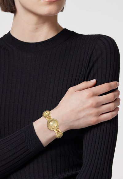 Auksinė apyrankė su apskritais žiedais, turinti tekstūruotą paviršių ir centriniame taške laikrodžio ciferblatą, dėvima ant rankos prie dryžuoto juodo megztinio.