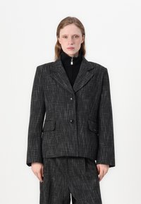Černý strukturální blazer s mírně tvarovaným střihem, dvěma předními kapsami, zapínáním na dva knoflíky a klopou s výřezem. V kombinaci s černými kalhotami.