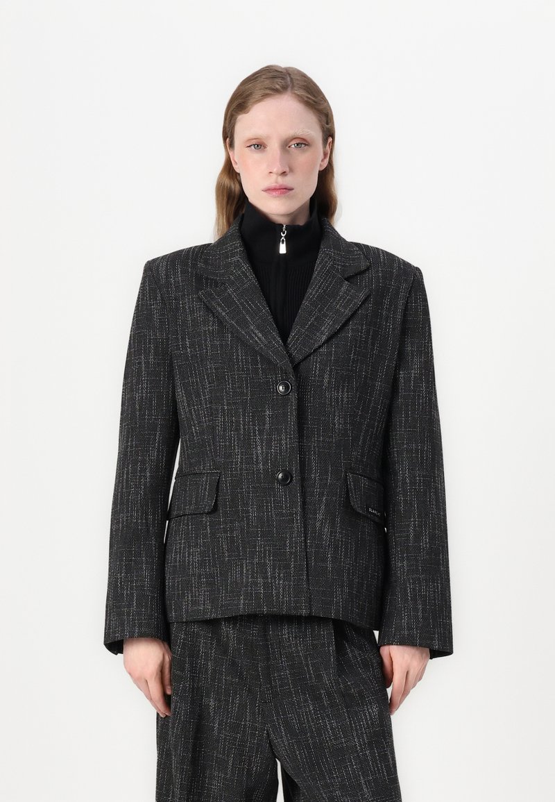 Černý strukturální blazer s mírně tvarovaným střihem, dvěma předními kapsami, zapínáním na dva knoflíky a klopou s výřezem. V kombinaci s černými kalhotami.