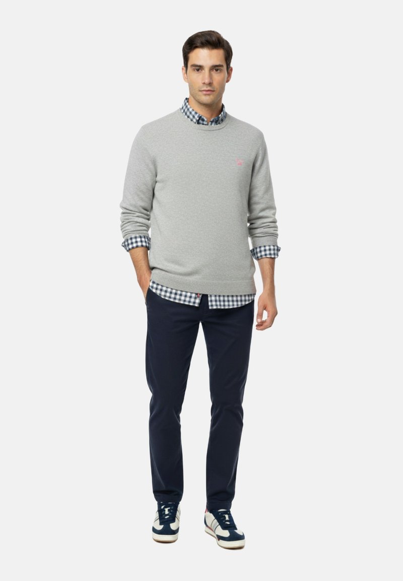 Suéter gris sobre una camisa de cuadros azul, pantalones azul marino y zapatillas azul marino. El suéter cuenta con un pequeño logo rosa. Tejido suave, corte relajado.