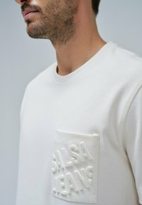 Bela dukserica s teksturiranim žepom na kojem je utisnut tekst "Salsa Jeans." Napravljena od mekog materijala, ima okrugli izrez i rebraste detalje.