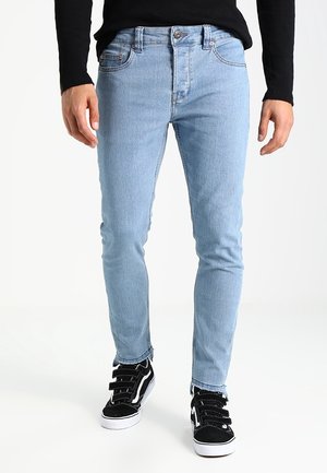 Personne portant un jean skinny bleu clair avec des ourlets effilochés et des baskets noires avec semelles blanches et bandes Velcro.