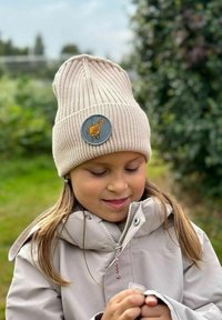 Väri beanie, jossa on ribbikuvio ja pyöreä merkki, jossa on keltainen lehmäkuvio. Lasten päällä, jotka on pukeutunut vaaleaan takkiin.
