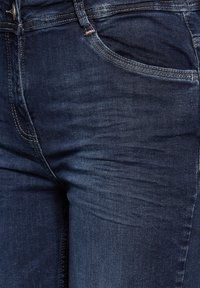 Pantalones vaqueros de mezclilla azul oscuro con corte ajustado, presentando bolsillos frontales, una cremallera y costuras en contraste a lo largo de las costuras.