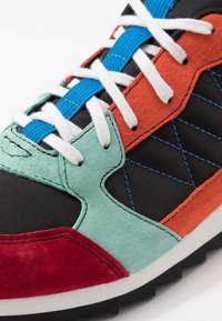 Flerfärgad sneaker med svart tyg, texturerade röda och gröna mockadetaljer, blå snören och en robust gummisula med mönster för grepp.