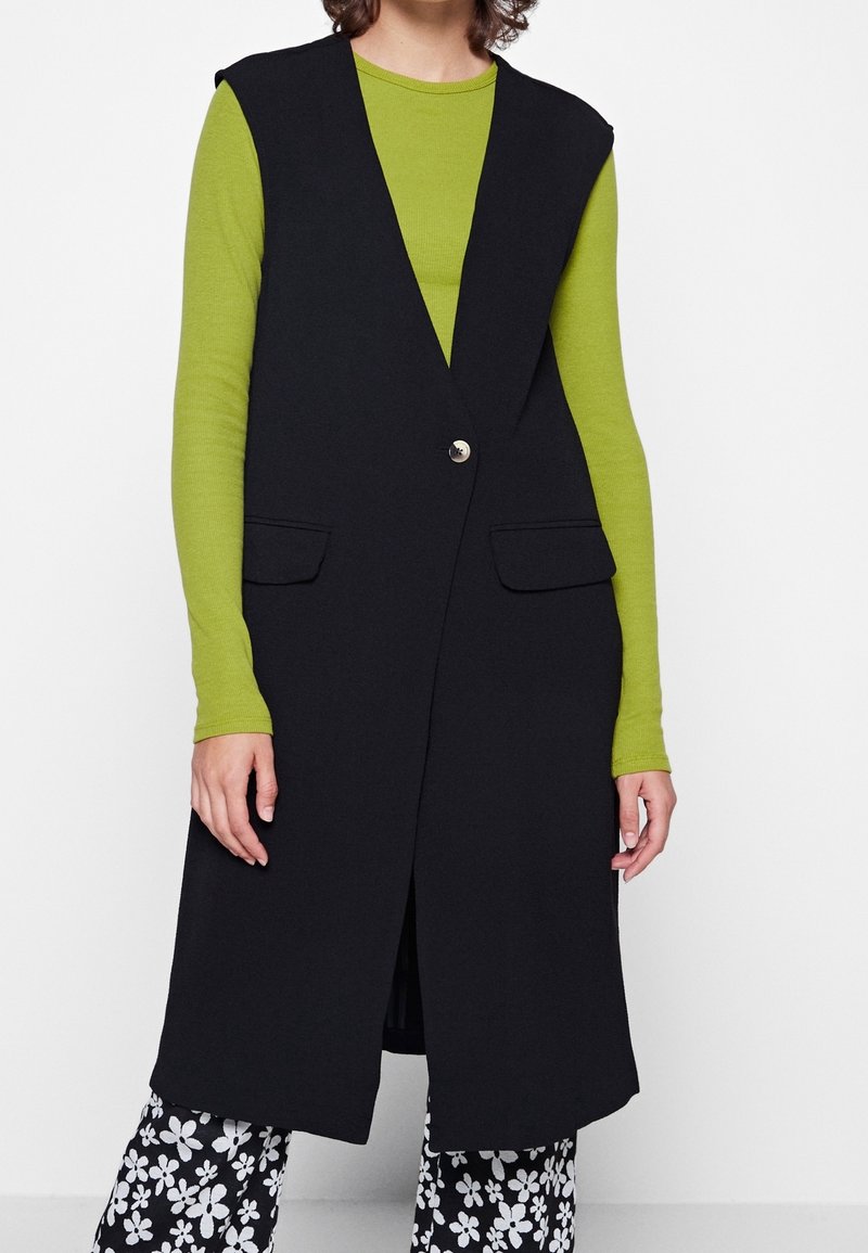 Svart ärmlös väst med djupt v-ringad hals, som har en enda knappstängning och två framfickor, lager på en grön långärmad topp.