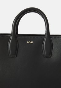 Čierna kožená kabelka s dvojitými uchami, ktorá sa vyznačuje hladkou textúrou a minimalistickým dizajnom. Obsahuje zlatý detail loga "BOSS".